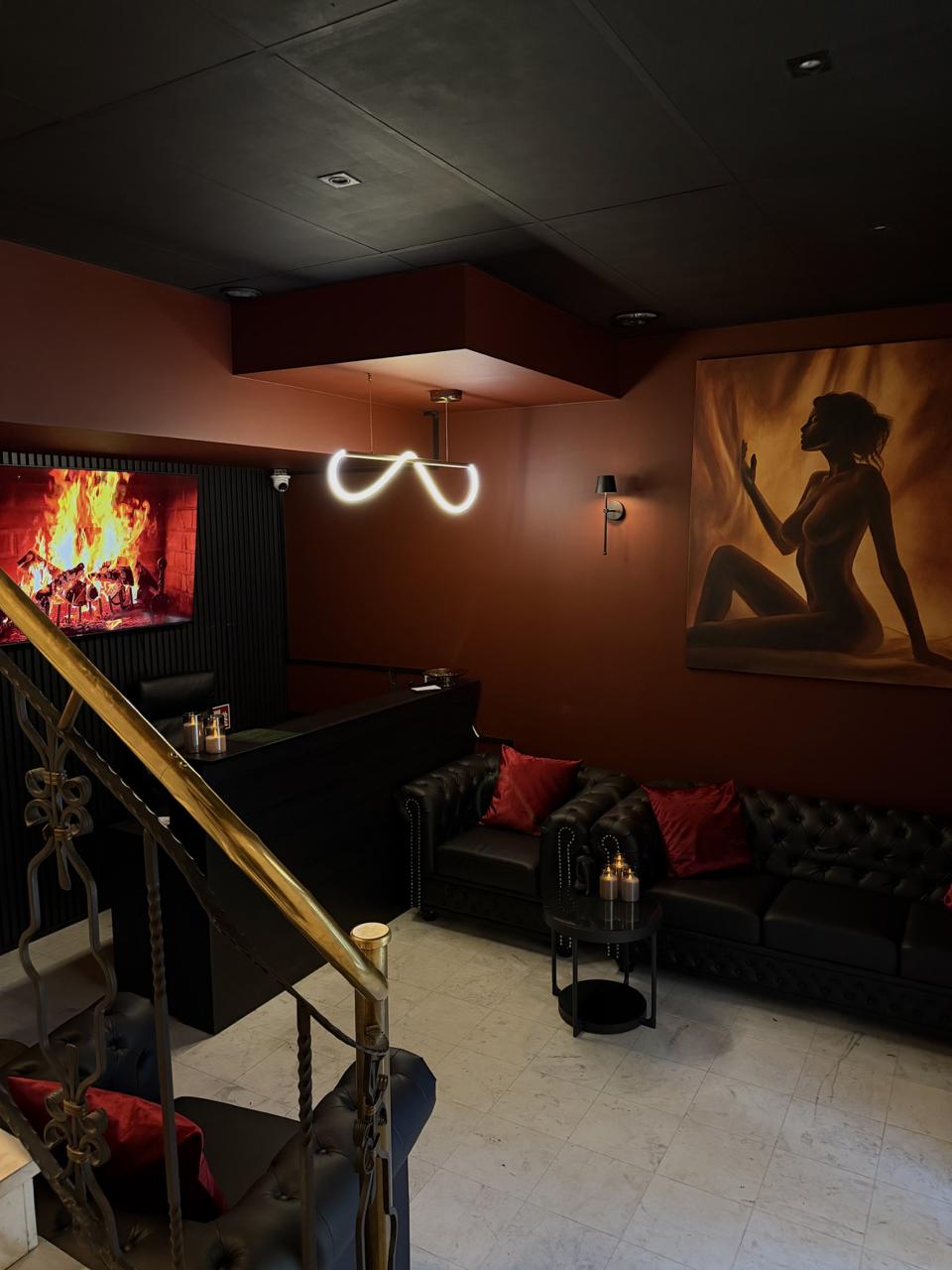 Recepcja i lounge UTOPIA: czarne sofy chesterfield z czerwonymi poduszkami, ogień na ekranie i świetlna pętla nad ladą